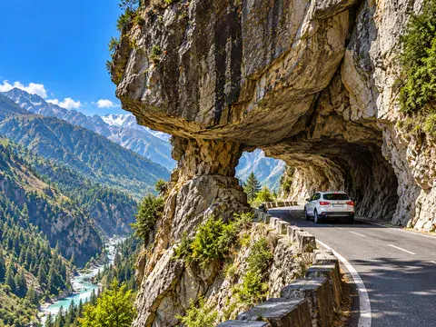 White vehicle driving through a carved rock cliff tunnel along a winding mountain highway above a river valley.